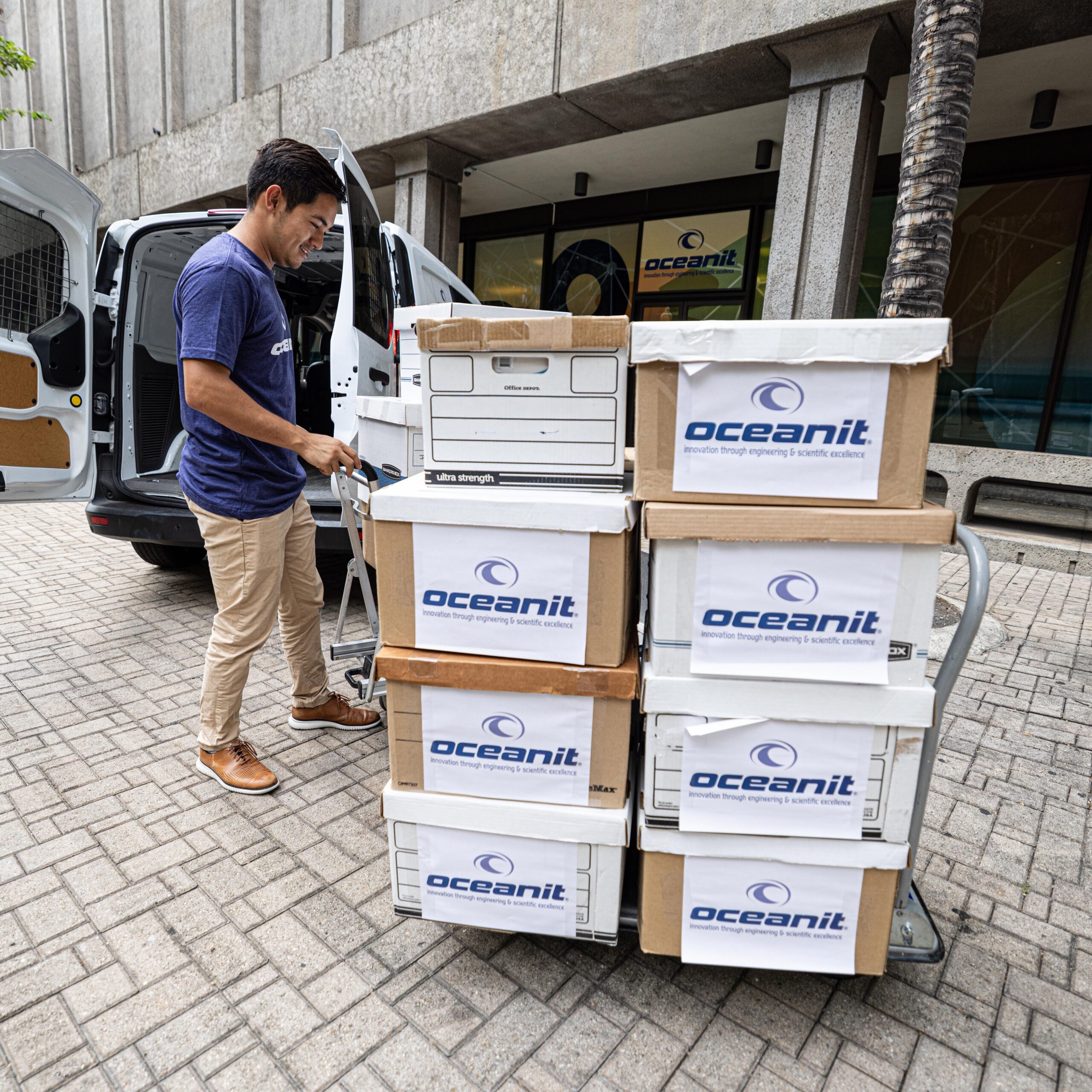 An employee maneuvering banker's boxes with the Oceanit logo into a sprinter van