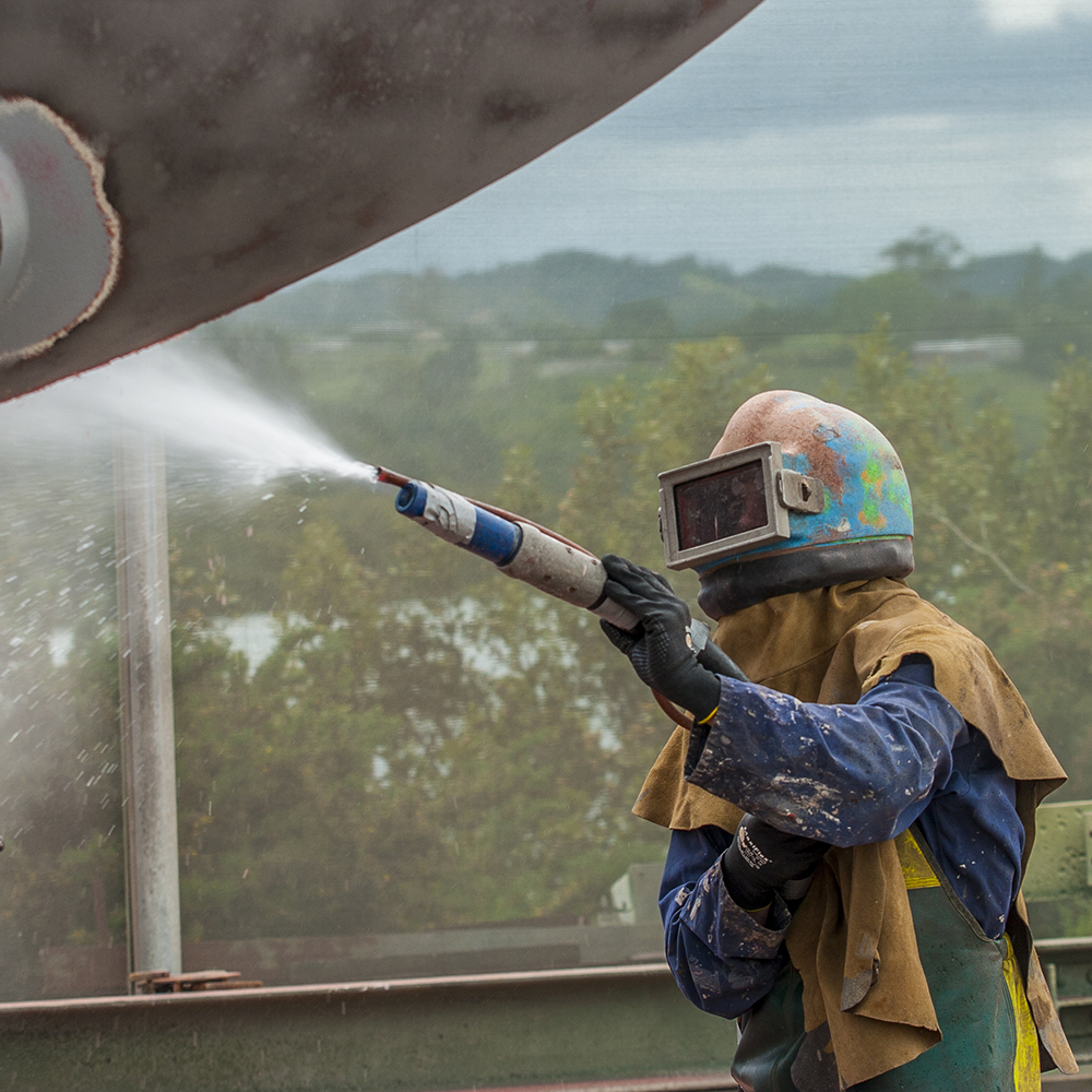 A worker in safety apparel sandblasting the corroded hull of a sailing vessel with a high pressure sandblasting nozzle