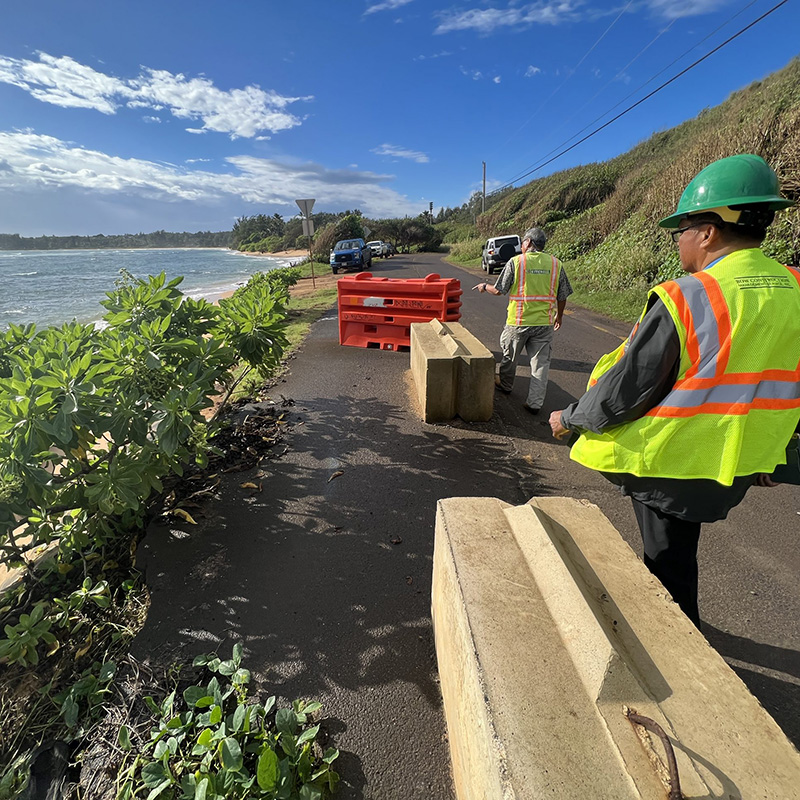 A road along Kauai's shore, with two engineers in safety vests pointing at concrete walls