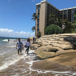 Half a dozen people walking along a shoreline and the sandbags along it, with a hotel in the background