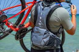 A man on the beach with his back to the camera lifting a bike and wearing a Honu pack