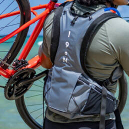 A man on the beach with his back to the camera lifting a bike and wearing a Honu pack