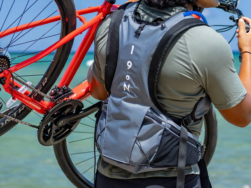 A man on the beach with his back to the camera lifting a bike and wearing a Honu pack