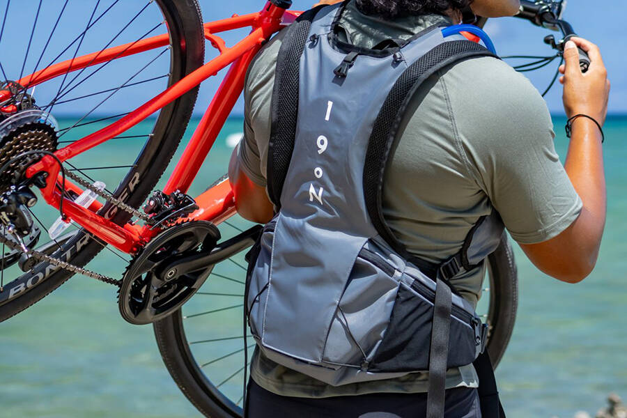 A man on the beach with his back to the camera lifting a bike and wearing a Honu pack