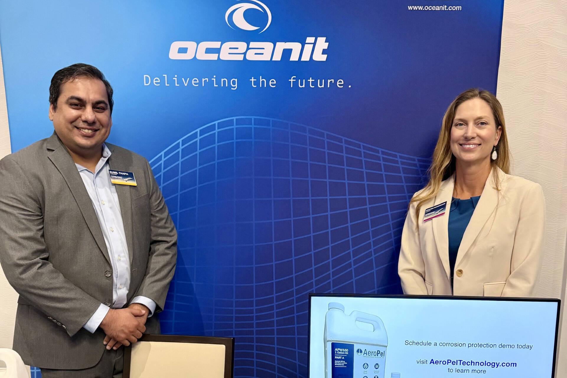 Two Oceanit employees standing in a booth with a blue Oceanit backdrop and an AeroPel jug
